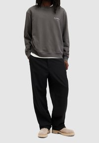 Grå crewneck sweatshirt med "ALLSAINTS"-logotyp, svarta löst sittande byxor och beige slip-on skor med tjock sula.