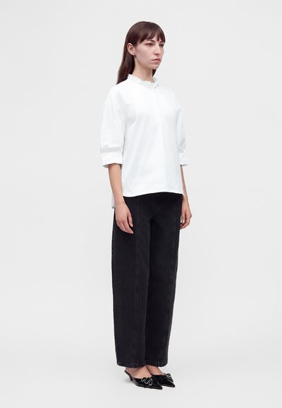Femme debout portant un chemisier blanc à manches trois-quarts, un pantalon noir à jambes larges et des chaussures noires à bouts pointus ornées de nœuds sur fond blanc.