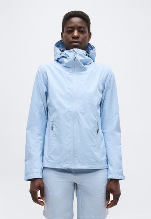 Άτομο που φοράει ανοιχτό μπλε ζακέτα με κουκούλα και φερμουάρ και ταίριασμα σορτς, στέκεται μπροστά σε λευκό απλό φόντο.