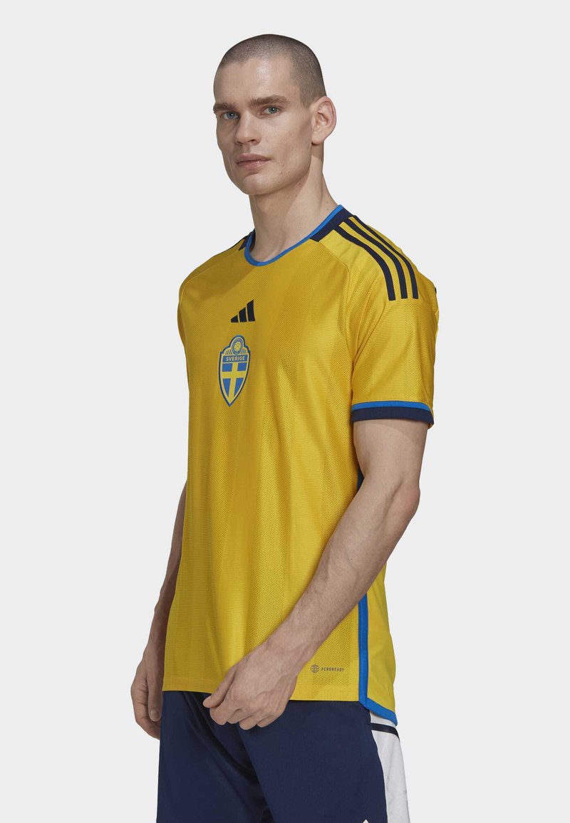 Camiseta de fútbol amarilla con acentos azules, que presenta un cuello redondo, mangas cortas y un escudo del equipo. Fabricada con material de malla transpirable.
