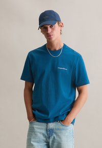 Turkusowa bawełniana koszulka z krótkimi rękawami i okrągłym dekoltem. Z subtelnym, białym haftowanym logo. Noszona z jasnoniebieskimi dżinsami i ciemną czapką.