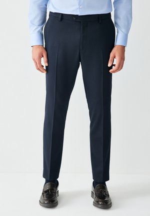 Mann trägt dunkelblaue Anzughose, hellblaues Knopfhemd und dunkelbraune Tassel-Loafer, steht vor weißem Hintergrund.