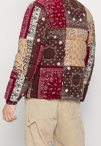 Vadderad jacka med en lapptäckedesign i burgundy, brunt och beige med bandana-mönster; elastiska manschetter; och en avslappnad passform.