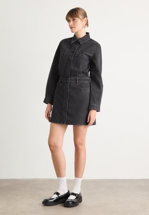 Abito camicia in denim nero con maniche lunghe, chiusura con bottoni, due tasche sul petto e vita con cintura. Indossato con scarpe nere e calzini bianchi.