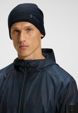 Navy knitted beanie with textured design and prominent "BOSS" branding on the side, worn with a dark patterned jacket.