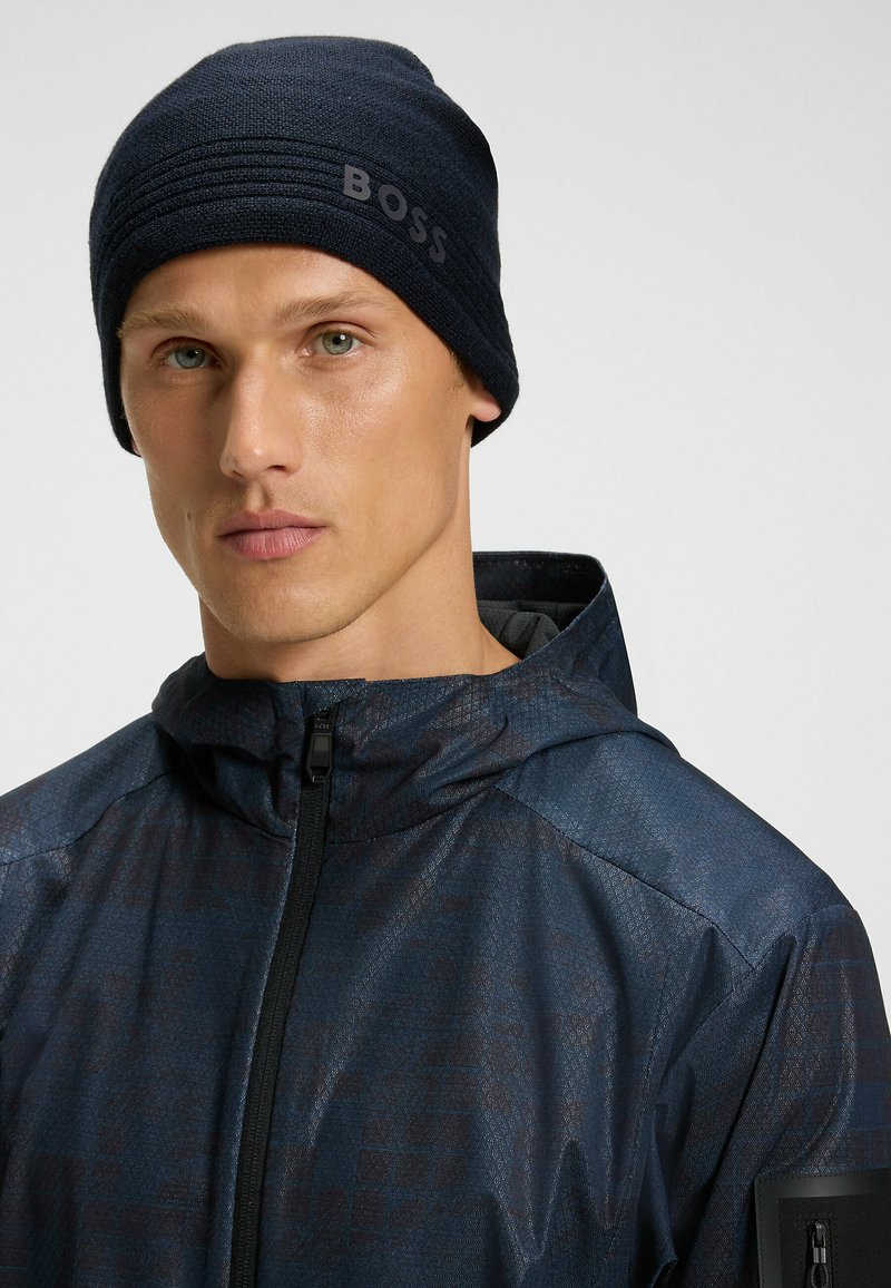 Berretto a maglia blu navy con design testurizzato e con il prominente marchio "BOSS" sul lato, indossato con una giacca scura a motivo.
