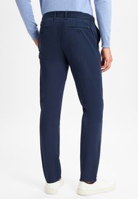 Navy pants with a tailored cut, flat front, and back pockets. Made of smooth fabric. Paired with white sneakers.
