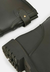 Tmavo zelené nepremokavé topánky z gumového materiálu, zaoblený špička, textúrovaná podrážka a popruh s detailom mosadzného gombíka.