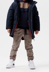 Doudoune marine avec accents rouges, hoodie superposé beige et marine, pantalon à carreaux rouge, noir et beige, et baskets blanches avec logo noir.
