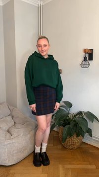 Sweatshirt verde oversize, mini saia de padrão xadrez azul-marinho, meias brancas, sapatos plataforma pretos. Em pé num chão de madeira ao lado de uma planta em vaso.