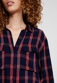 Navy blue plaid shirt with red and white stripes, cotton fabric, button-up front, two chest pockets, and classic collar design.