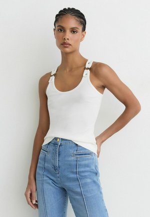 Kvinde med flettet hår iført en hvid tanktop med spænder på stropperne og højtaljede blå jeans, stående med den ene hånd på hoften.