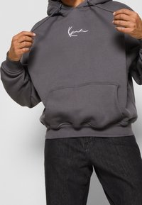 Grijze hoodie van zacht materiaal, voorzien van een voorzak en een wit logo op de borst. Voorzien van gladde trekkoordjes en geribbelde manchetten.