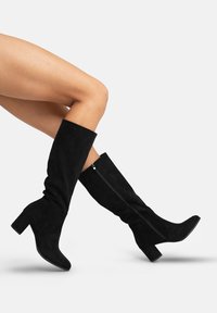 Botas altas de piel de ante negra con tacón de bloque y cremallera lateral, que presentan un diseño arrugado y una textura suave.