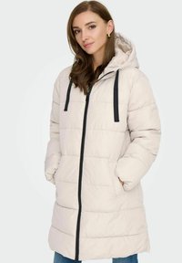 Langer beiger Puffer-Mantel mit Kapuze, schwarzem Reißverschluss und Kordelzug-Akzenten. Dicke gestepfte Textur und Seitentaschen.