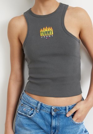 Person iført grå ærmeløs cropped top med "Tommy Jeans"-logo og flammedesign, kombineret med blå cowboybukser med hånden i lommen.