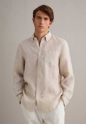 Junger Mann mit braunen Haaren, der ein beiges Hemd mit Knöpfen und weiße Hosen trägt und mit den Händen in den Taschen vor einem schlichten Hintergrund steht.