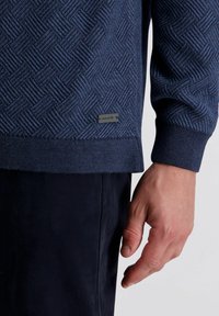 Marineblaues Sweatshirt mit einem strukturierten Rautenmuster, gerippten Bündchen und Saum. Verfügt über ein kleines Logotag in der unteren linken Ecke. Weiches Strickmaterial.
