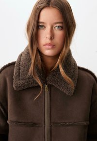 Giacca in shearling marrone con colletto in pelliccia spessa, chiusura con zip e due tasche frontali. Il tessuto ha una texture morbida e una finitura liscia.