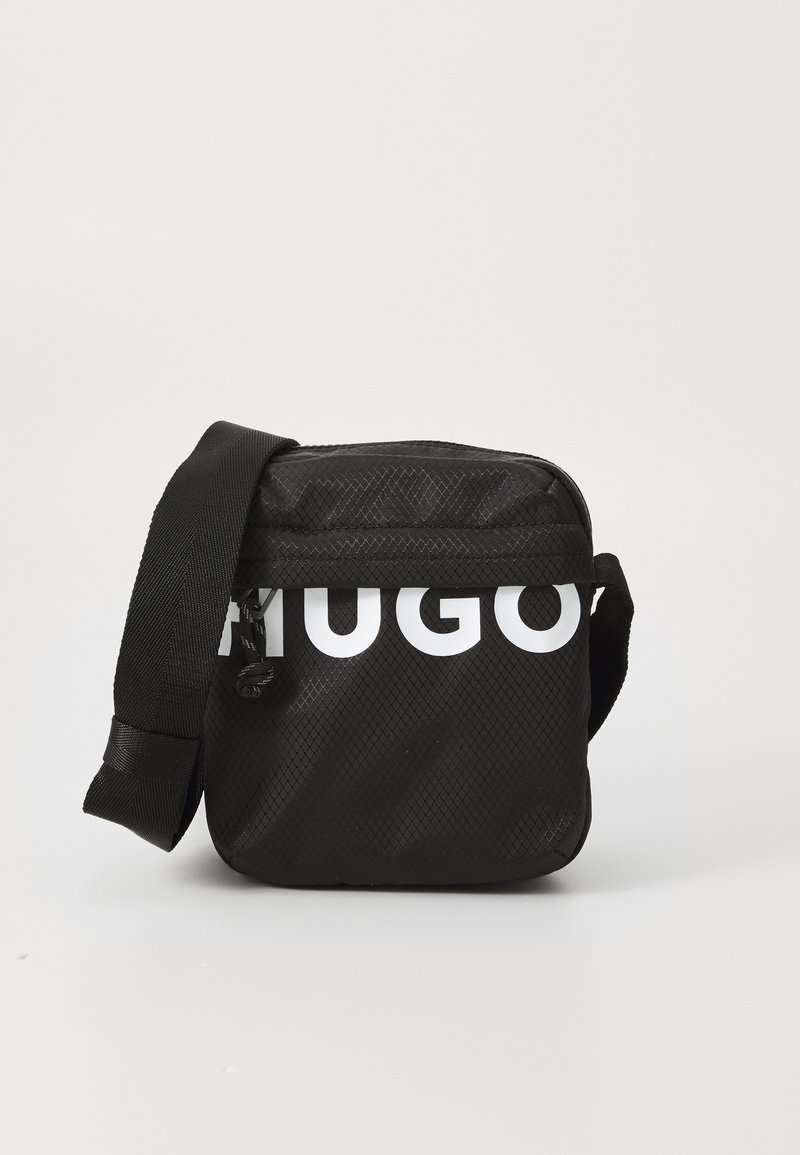 Bolso negro de bandolera con una superficie texturizada, que presenta un logo blanco. Incluye una correa ajustable y cierre con cremallera. Forma cuadrada compacta.