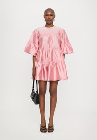 Ροζ σατέν κοντό φόρεμα με φουσκωτά μανίκια και σχεδίαση με ριγέ φραμπαλά. Συνδυασμένο με μαύρη τσάντα και σάνταλ με λουριά. Απλή φόντο.