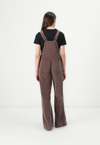 Pettorina in corduroy marrone con gambe larghe, dotata di due tasche posteriori, bretelle regolabili e texture a righe verticali. Camicia nera sottostante.