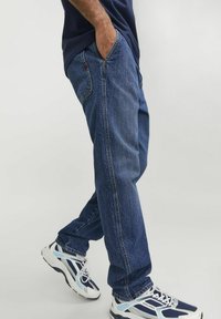 Jeans in denim blu con una vestibilità rilassata, dotati di tasche e cuciture a vista. Abbinati a scarpe da ginnastica bianche e blu.