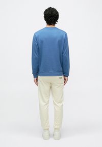 Zils kokvilnas džemperis ar apaļu kaklu, ribotām aprocēm un bēša krāsas kokvilnas sporta bikses ar elastīgu vidukli un logo aizmugurējā kabatā. Baltas sporta kurpes.