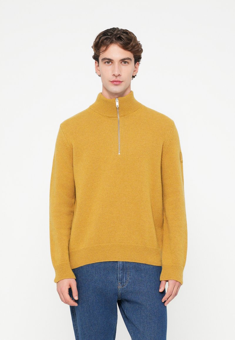 Sárga cipzáros pulóver magas nyakkal, bordás mandzsettákkal és szegéllyel. Puha textúra, laza fazon. Kék farmernadrággal viselve.