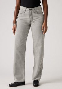 Personne portant un jean gris clair droit, des chaussures noires et un haut noir sans manches, debout devant un fond uni.