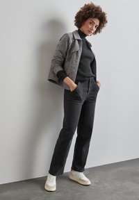 Grå texturerad jacka över en svart ribbad polotröja, i kombination med höga svarta jeans och vita sneakers. Modellen poserar med händerna i fickorna.