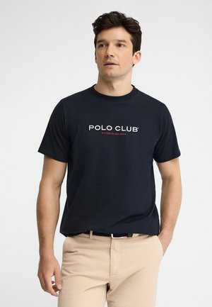 Junger Mann steht, trägt ein schwarzes „Polo Club“-T-Shirt und beige Hose, mit einer Hand lässig in der Tasche vor einem einfarbigen Hintergrund.