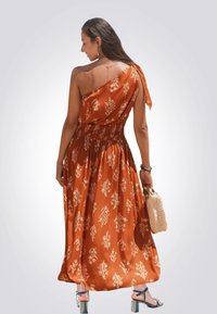 Ein schulterfreies, fließendes Maxikleid in verbranntem Orange mit weißen Blumenmustern, gerafftem Bund und einem seitlichen Bindeband. Kombiniert mit einer Häkel-Tasche.