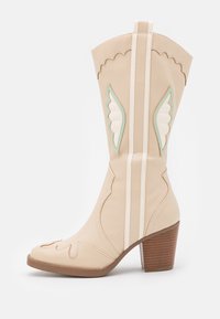 Bota de vaquero beige hecha de cuero suave con patrones decorativos de alas en blanco y azul claro, que presenta un tacón de madera apilada.