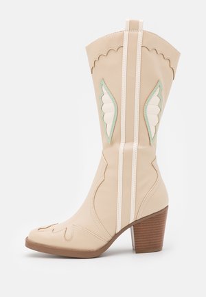 Bota de vaquero beige hecha de cuero suave con patrones decorativos de alas en blanco y azul claro, que presenta un tacón de madera apilada.