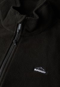 Giacca in pile nera con chiusura a mezza zip, colletto alto e piccolo logo ricamato sul lato sinistro. Texture morbida e design leggero.