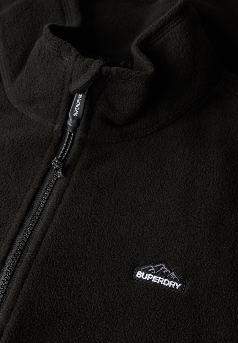 Veste en polaire noire avec fermeture à demi-glissière, col haut et petit logo brodé sur le côté gauche. Texture douce et design léger.