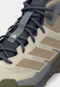 Beiger und grauer Sportschuh mit strukturierter Oberfläche, Mesh-Details und grünen Akzenten. Verfügt über graue Schnürsenkel und drei weiße Streifen an der Seite.