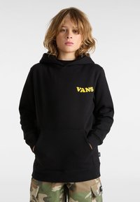 Felpa nera in misto cotone con tasca frontale e logo "VANS" giallo. Abbinata a pantaloni cargo mimetici con tasche laterali e vestibilità rilassata.