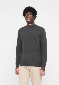 Pull en maille gris foncé avec un col rond, des manches longues et un petit logo jaune sur la poitrine. Il présente une texture douce et un design simple.