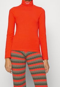 Långärmad polokrage i klar orange bomull med en jämn textur. Kombinerad med mönstrade leggings i gröna, röda och grå zigzags.