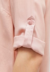 Camicia button-up rosa chiaro con maniche, caratterizzata da un polsino arrotolato, dotata di un bottone rosa e una trama di tessuto liscia.