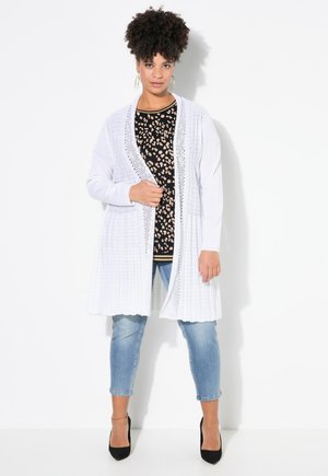 Cardigan bianco lungo con apertura frontale, texture lavorata a maglia e dettagli decorativi. Abbinato a un top con stampa leopardata e jeans azzurri chiari. Tacchi eleganti neri.