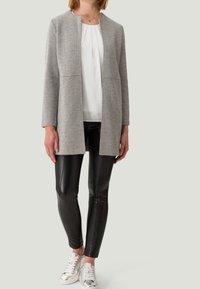 Manteau gris texturé à coupe droite, associé à des leggings noirs brillants et des baskets argentées. Chemisier blanc en dessous. Design simple et moderne.