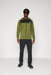 Mann iført svarte solbriller, olivengrønn og svart jakke med glidelås, svarte bukser og svarte sko, står mot en ensfarget bakgrunn.