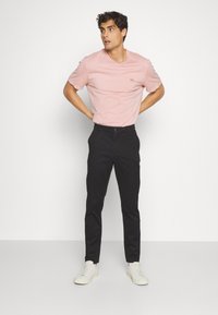 T-shirt di cotone rosa chiaro con un piccolo logo ricamato, abbinata a pantaloni neri slim-fit e sneakers bianche, in piedi contro uno sfondo neutro.