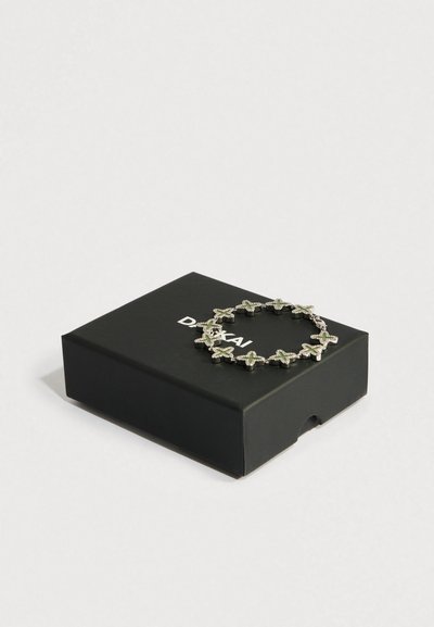 Pulseira em tom prateado com formas de trevo entrelaçadas adornadas com cristais verdes e transparentes, repousando sobre uma caixa preta com texto branco.