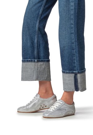 Chaussures de sport argentées à texture perforée, lacets blancs, accents latéraux prononcés, portées avec un jean bleu foncé retroussé et des revers bleu clair.