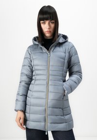 Vrouw in een lichtblauwe gewatteerde winterjas met een capuchon en een ritssluiting aan de voorkant, staande tegen een effen witte achtergrond.