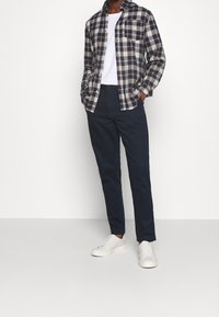 Koszula w kratę w kolorze granatowym i beżowym, biała koszulka, granatowe spodnie i białe sneakersy. Luźny krój z detalami w postaci guzików i kieszonką na piersi.